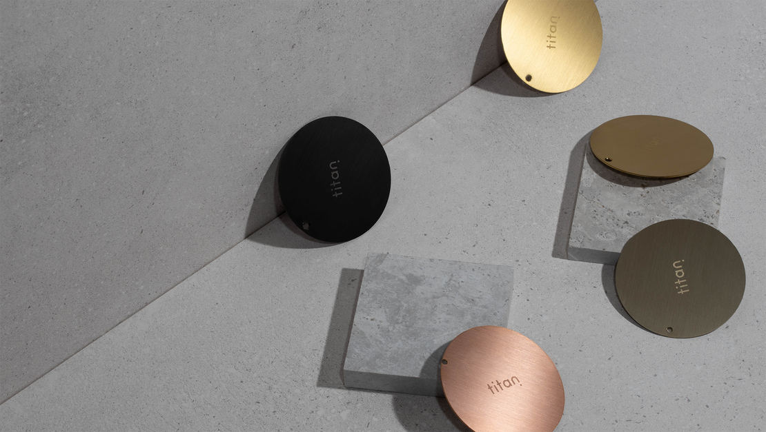 Still life photo of Titan finish samples in matte black, royal gold, rose gold, and pearl gold, arranged on a gray background with complementary gray set props, showcasing the variety of available finish samples.